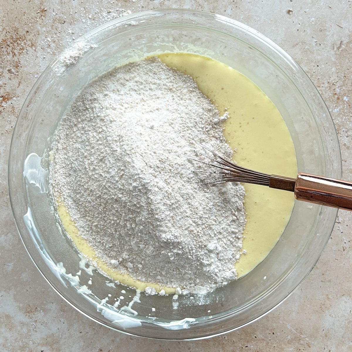A glass bowl with flour and dry ingredients on top of yellow batter, and a whisk resting inside, ready for mixing on a light countertop.