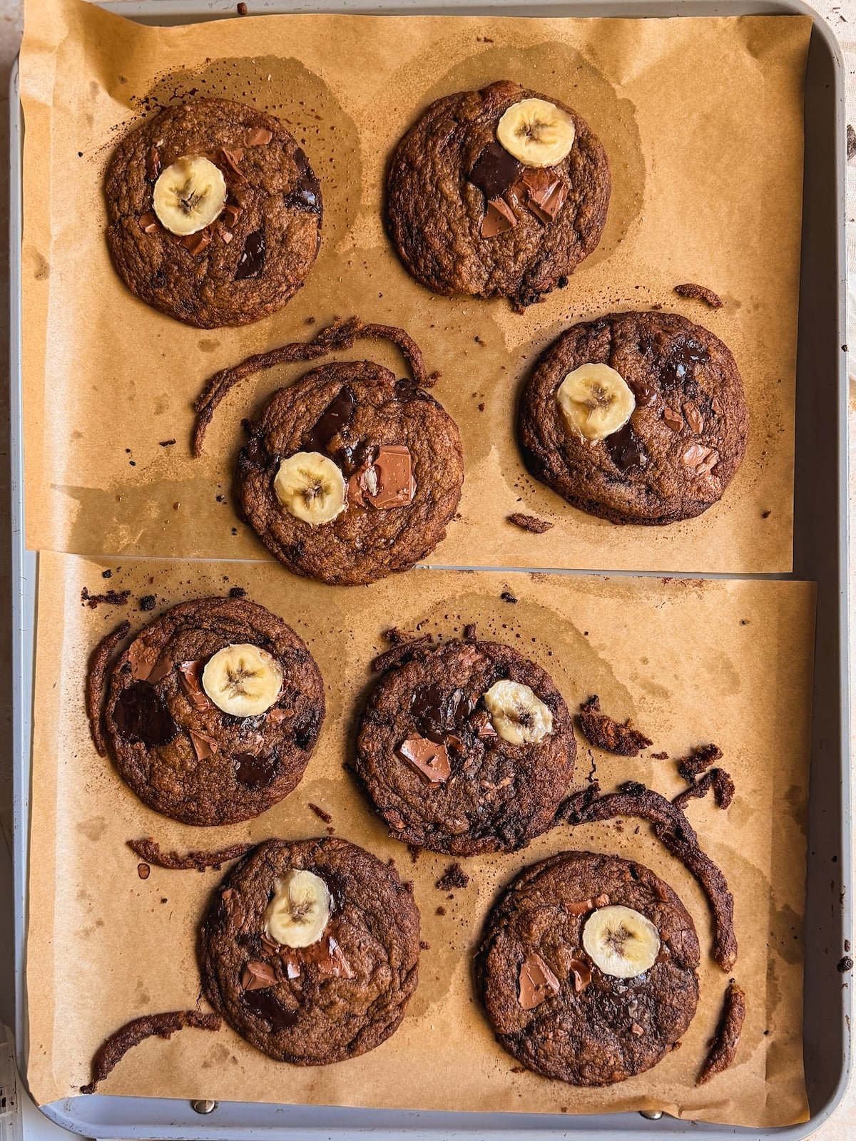 Eight chocolate chip banana bread cookies with banana slices on top, baked on parchment paper in a baking tray.