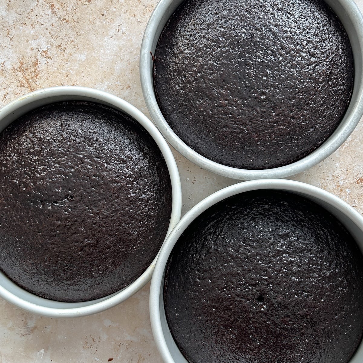 Three round chocolate cakes in white pans are arranged close together on a light, textured surface. The cakes have a smooth, dark surface and appear freshly baked.