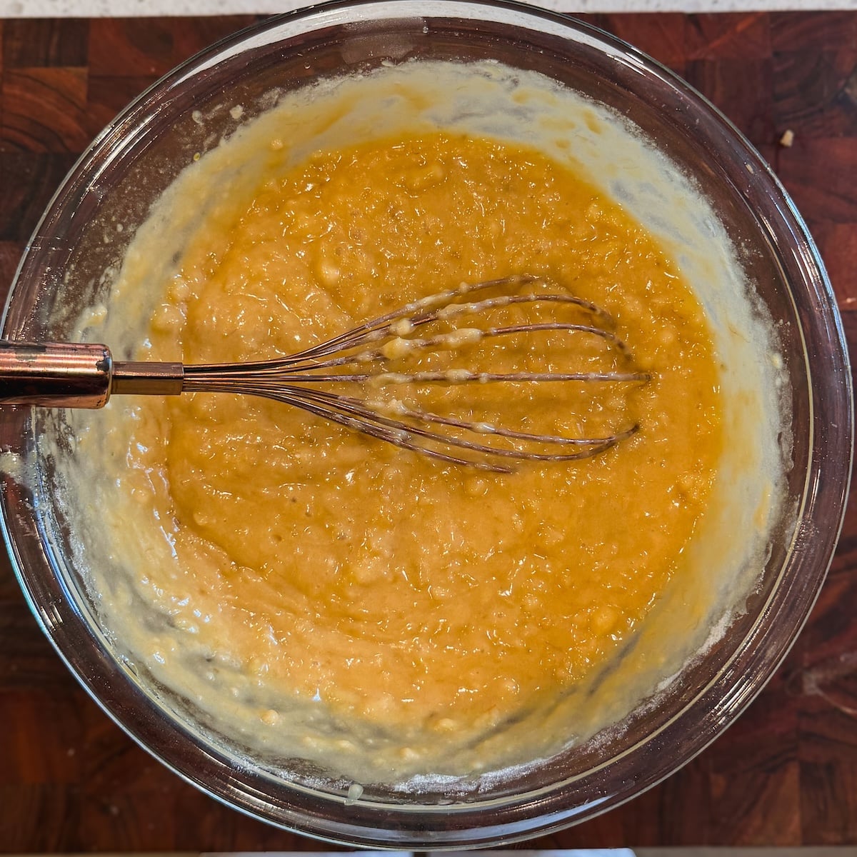 A glass bowl of yellow banana bread batter with a metal whisk rests on a wooden surface. The mixture appears thick and lumpy, with some batter clinging to the sides of the bowl.