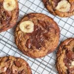 brown butter banana bread chocolate chip cookies with banana slices on top cooling on a wire rack. The cookies are golden brown with melted chocolate and sprinkled with sea salt.