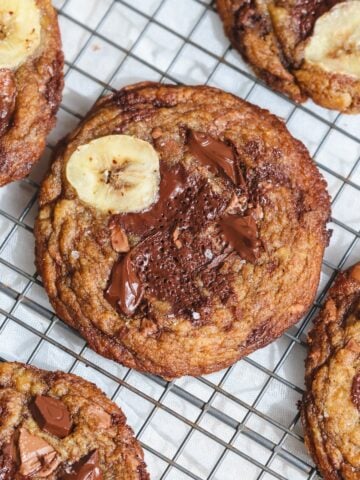 brown butter banana bread chocolate chip cookies with banana slices on top cooling on a wire rack. The cookies are golden brown with melted chocolate and sprinkled with sea salt.