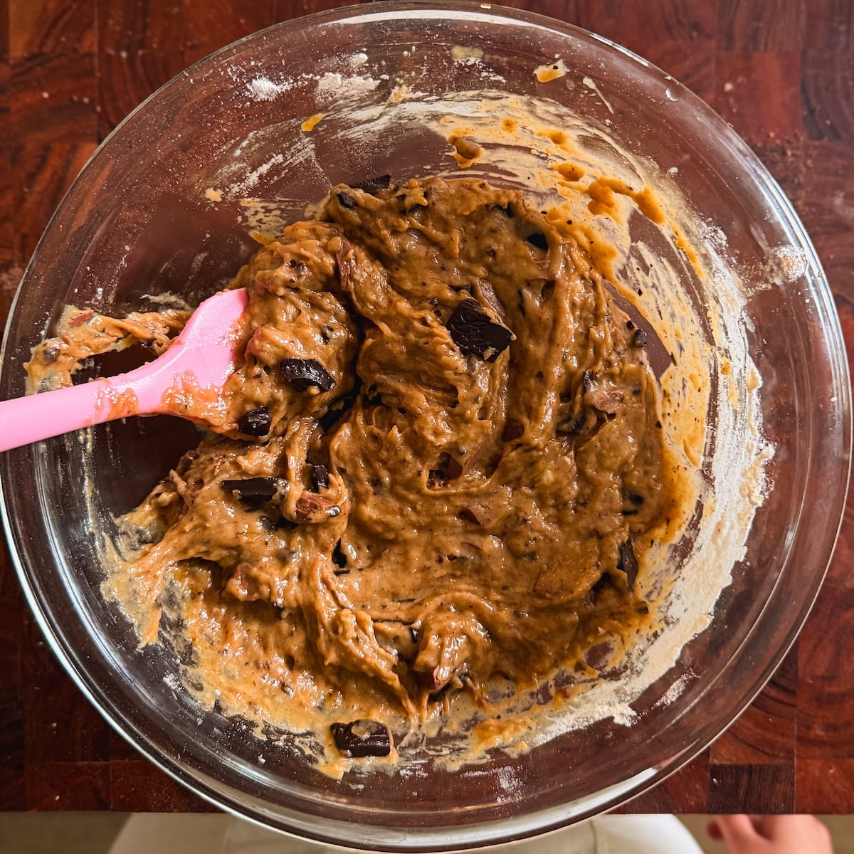 A glass bowl filled with banana bread chocolate chip cookie dough sits on a wooden surface. A pink spatula rests inside the bowl, partially covered in dough.