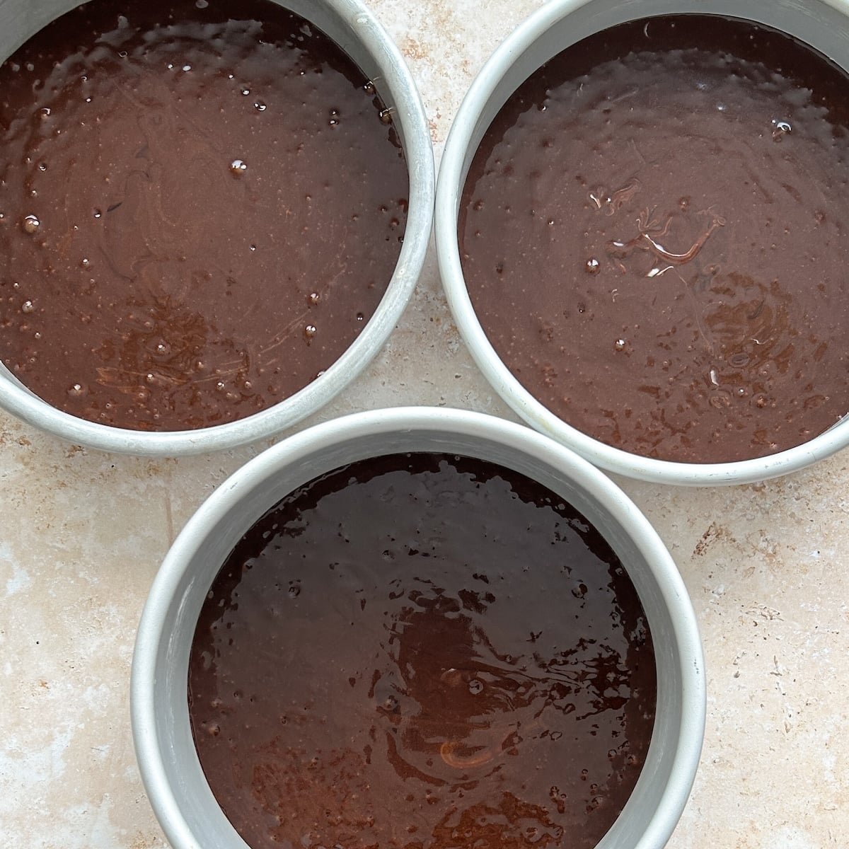 Three round cake pans filled with chocolate cake batter are arranged on a light-colored countertop, ready to be baked.
