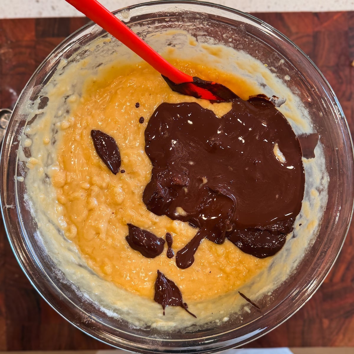 A glass bowl with banana bread batter and cocoa powder mixture on top, being mixed with a red spatula, sitting on a wooden surface.