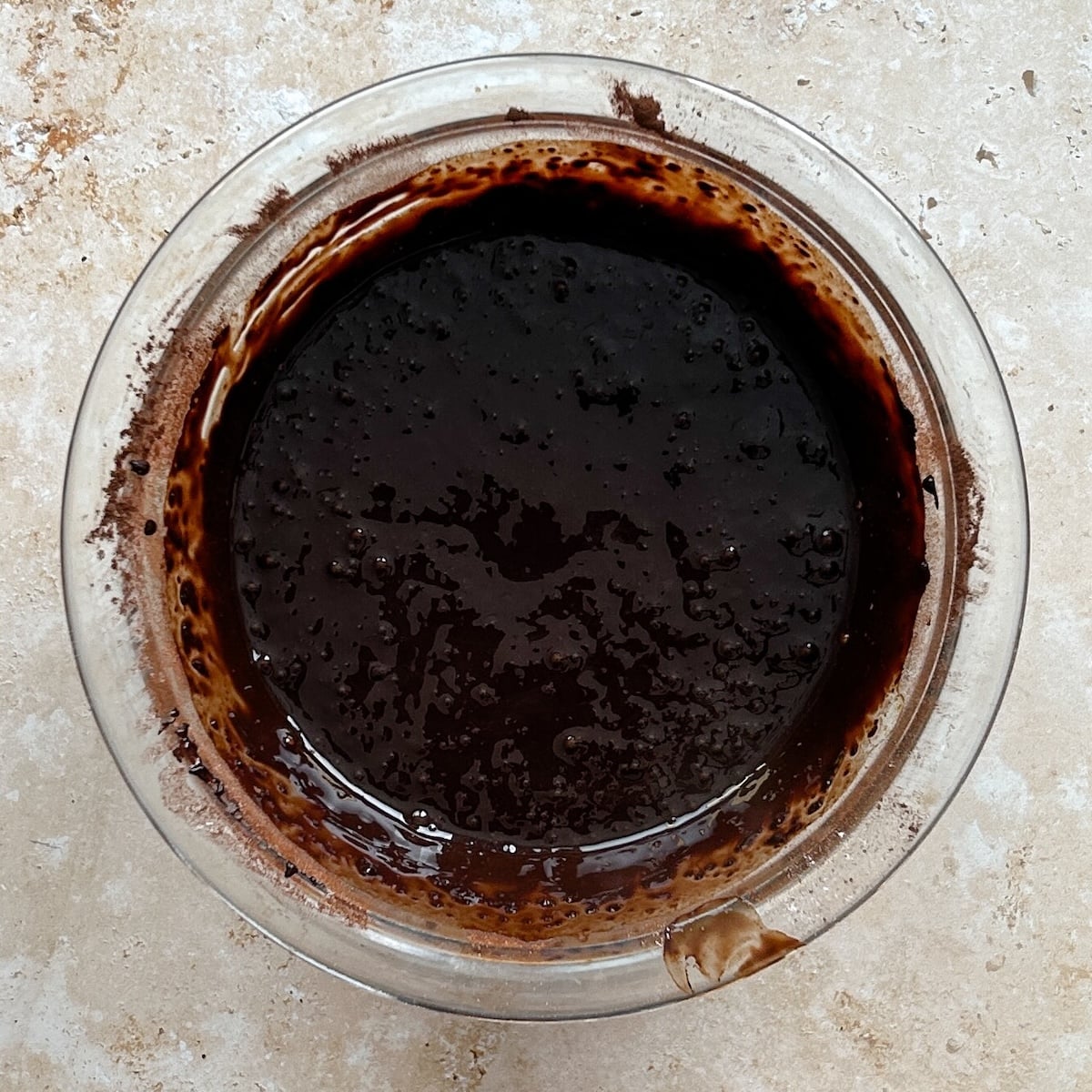 A clear glass bowl filled with glossy, dark chocolate mixture sits on a light-colored countertop. Some chocolate residue is visible on the rim and side of the bowl.