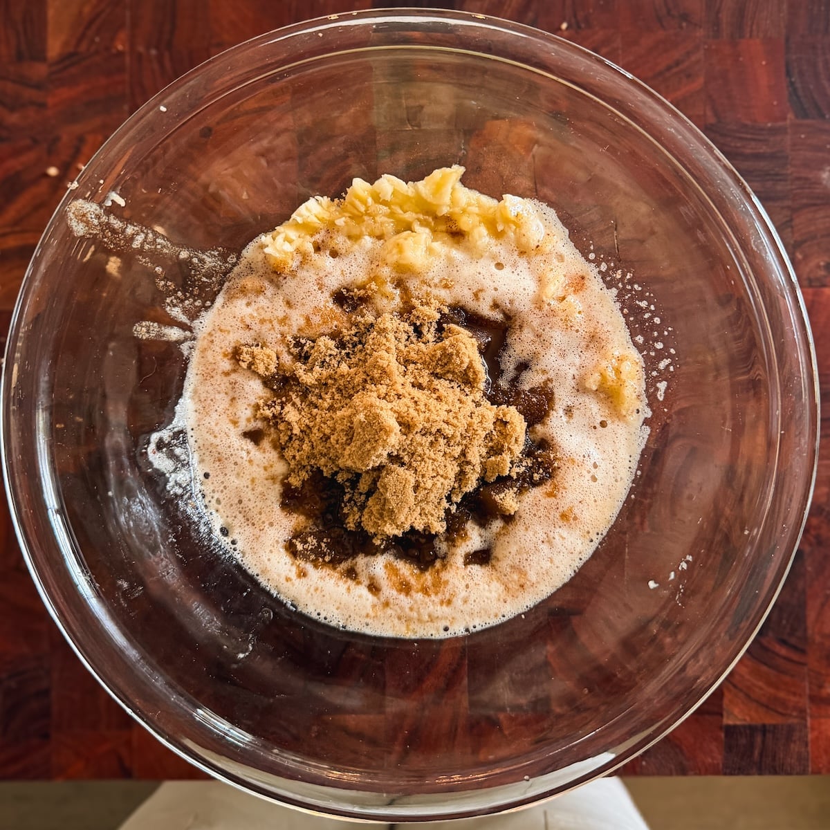 A glass bowl on a wooden surface contains mashed bananas, browned butter, and brown sugar, ready to be mixed