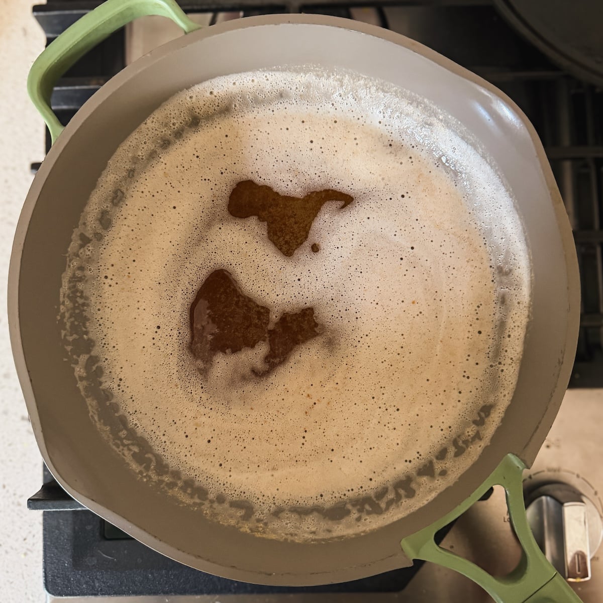 A light gray skillet with green handles is on a stove, containing simmering brown butter. Foam surrounds darker brown spots in the center of the bubbling liquid.