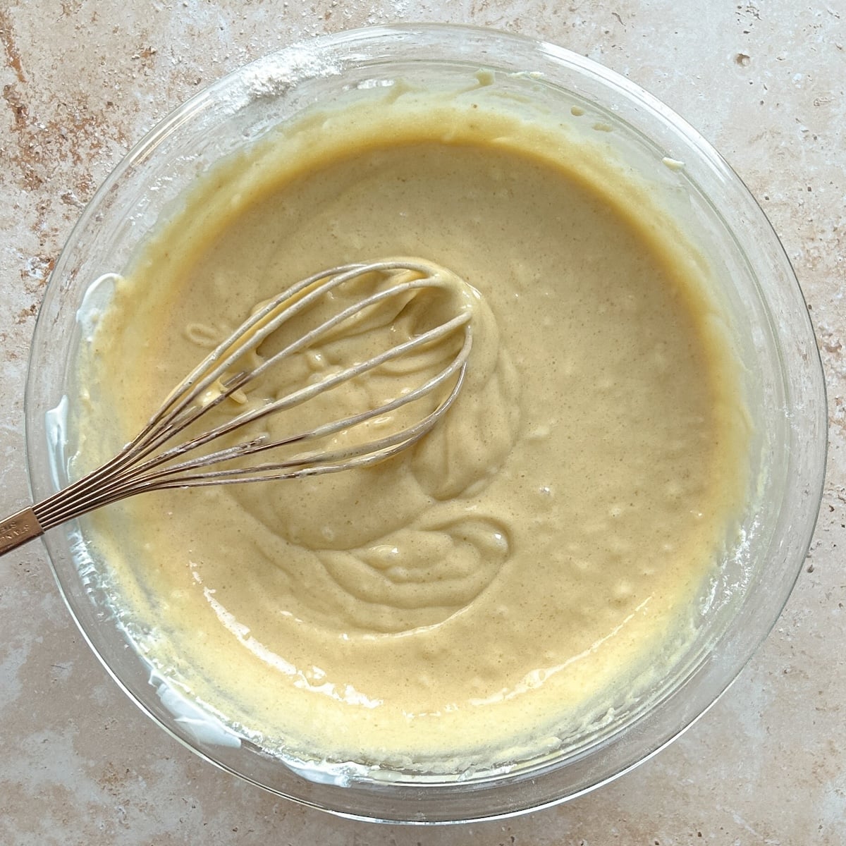A glass bowl filled with smooth, yellow cake batter and a metal whisk resting inside, on a light-colored countertop.
