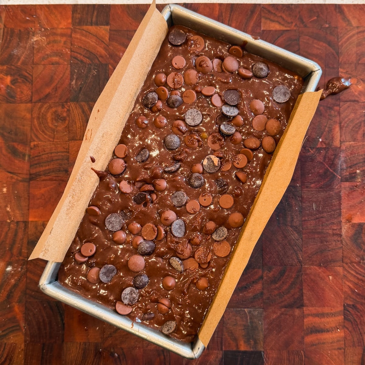 A loaf pan lined with parchment paper is filled with chocolate banana bread batter and topped with chocolate chips, resting on a wooden cutting board.