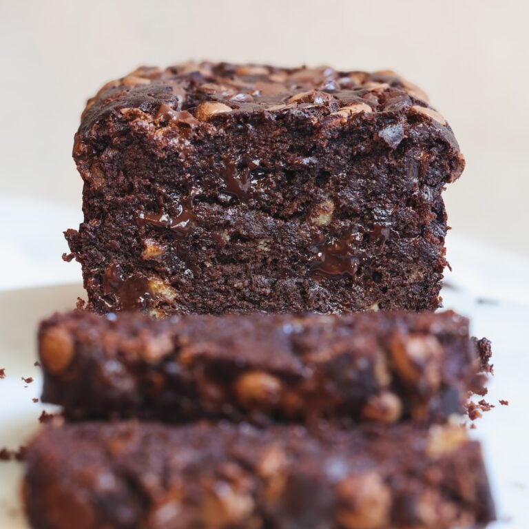A close-up of a thick slice of moist chocolate banana bread with melted chocolate chunks, resting on a white surface. Two out-of-focus slices lie in front of it.