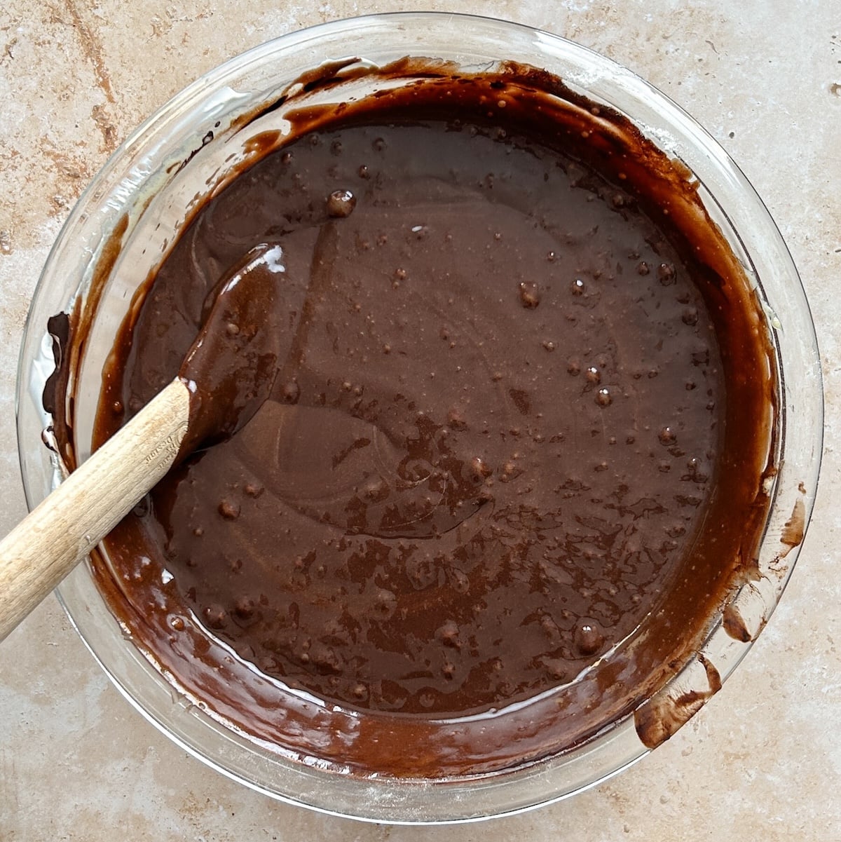 A glass bowl filled with chocolate cake batter sits on a light countertop, with a wooden spoon resting inside the bowl.