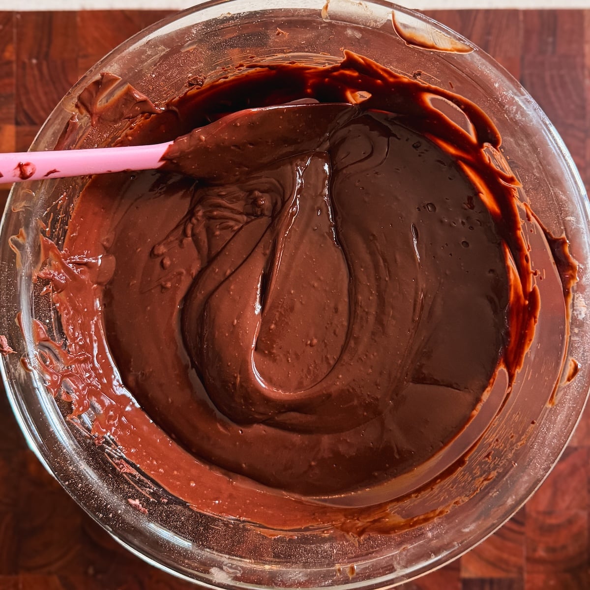 A glass bowl filled with smooth, glossy chocolate frosting is being mixed with a pink spatula on a wooden surface.