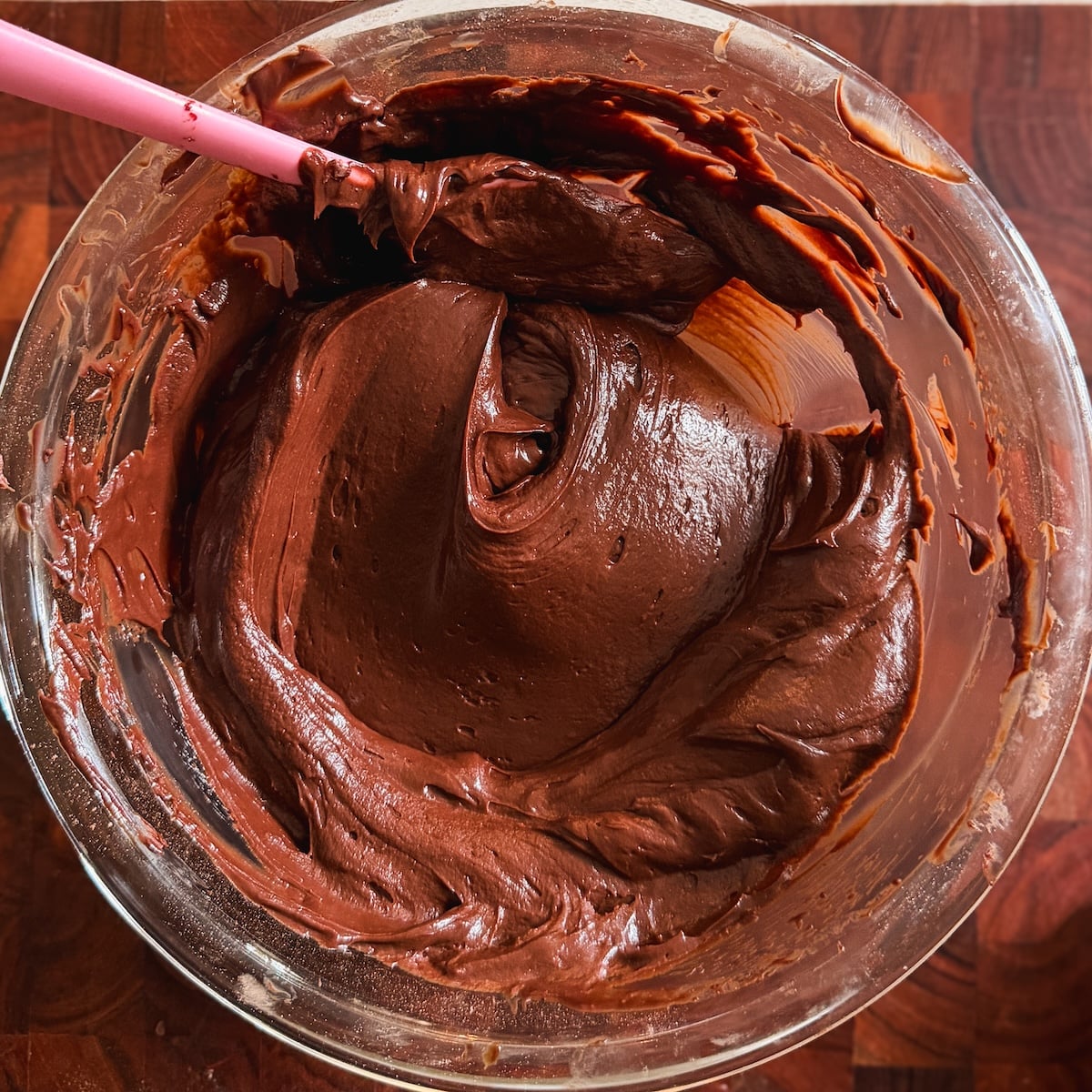 A glass bowl filled with smooth, rich chocolate frosting being mixed with a pink spatula on a dark wooden surface.