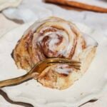 A cinnamon roll with white icing sits on a cream-colored plate. A gold fork rests in the roll, ready for a bite. A cinnamon stick is visible in the background. The roll looks soft and gooey with a cinnamon filling