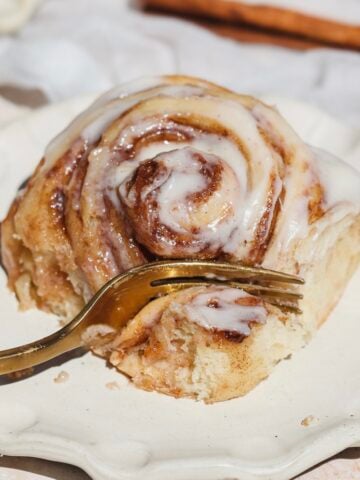 A cinnamon roll with white icing sits on a cream-colored plate. A gold fork rests in the roll, ready for a bite. A cinnamon stick is visible in the background. The roll looks soft and gooey with a cinnamon filling