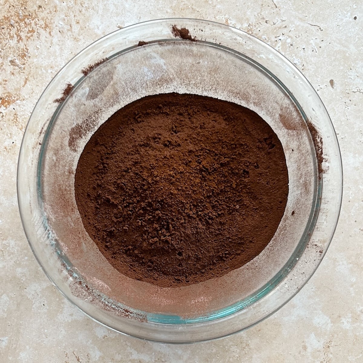 A glass bowl filled with a mound of finely ground dark brown cocoa powder sits on a light beige textured surface.