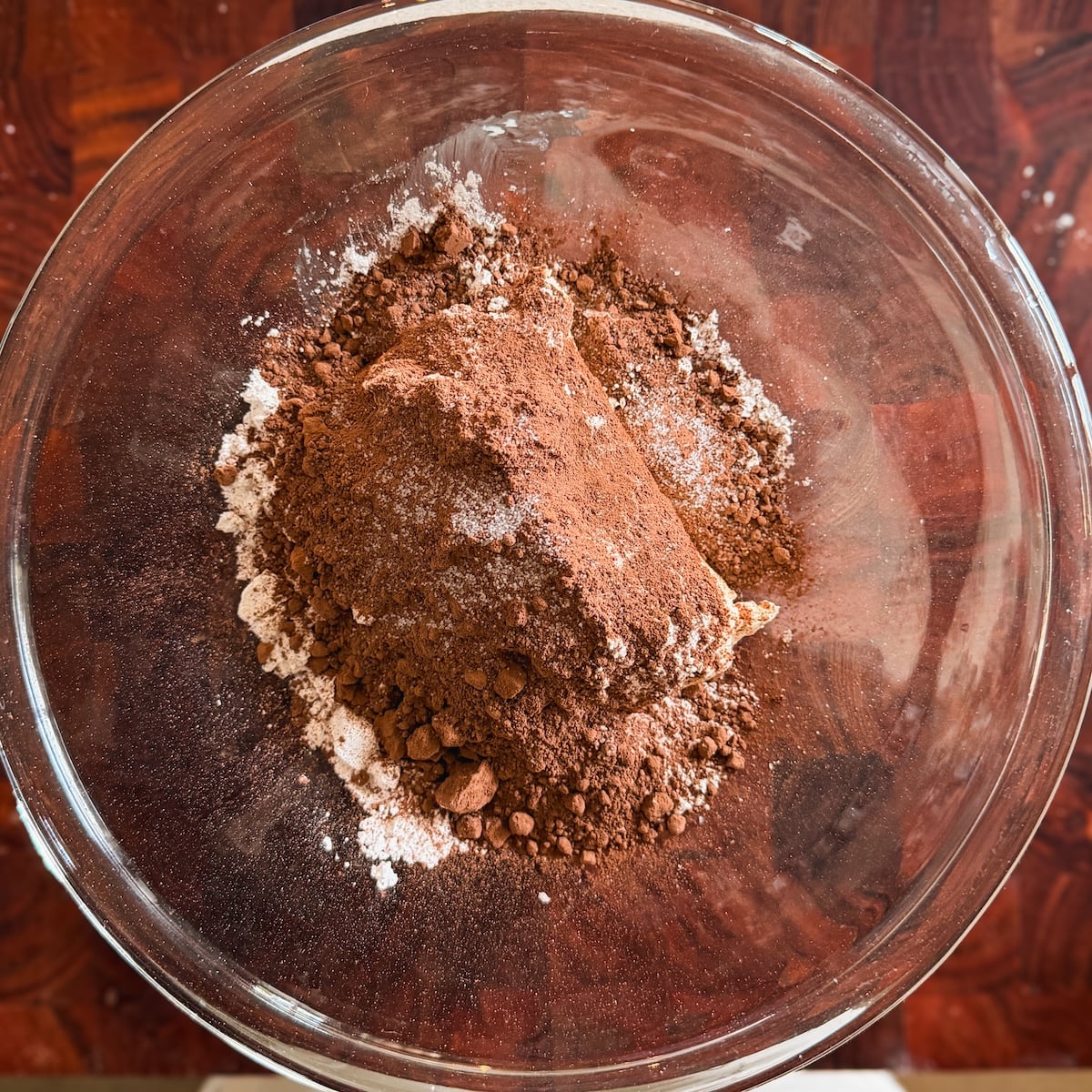 A glass bowl contains a pile of cocoa powder mixed with powdered sugar and a sprinkle of salt, sitting on a wooden surface