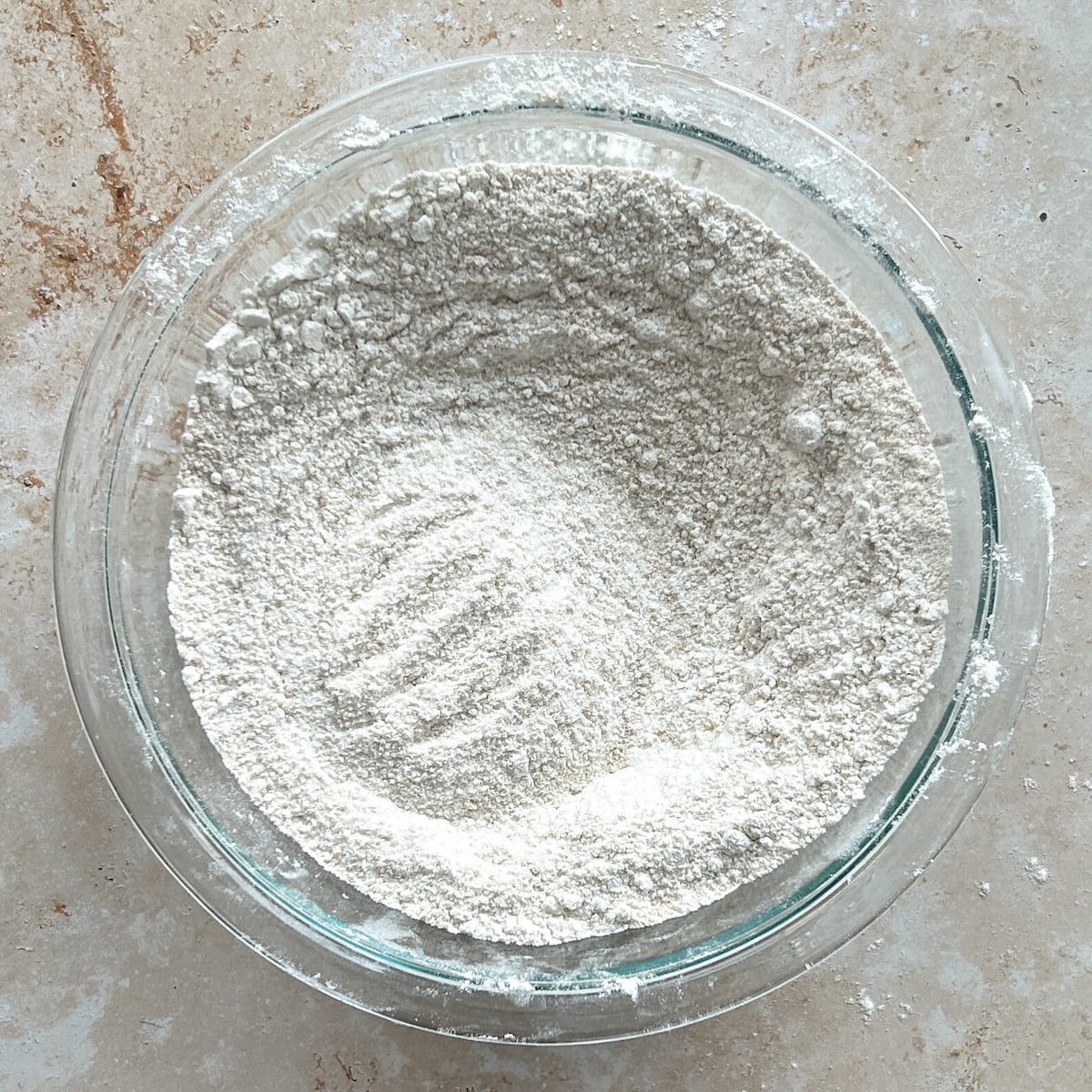 A glass bowl filled with white dry ingredients sits on a light brown countertop. Some finger marks are visible on the surface of the flour.
