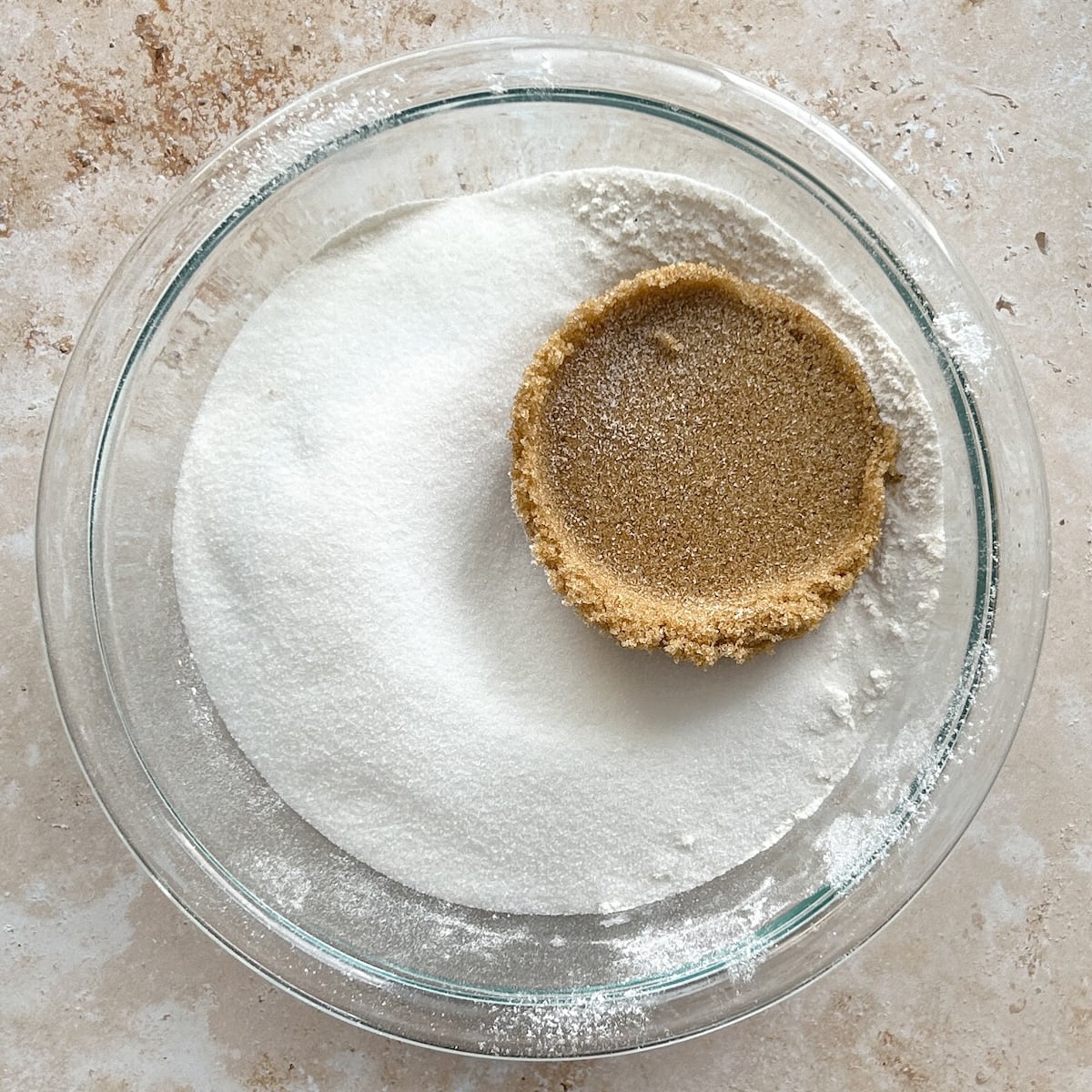 A glass bowl containing flour, white granulated sugar, brown sugar, and flour sitting on a light countertop.