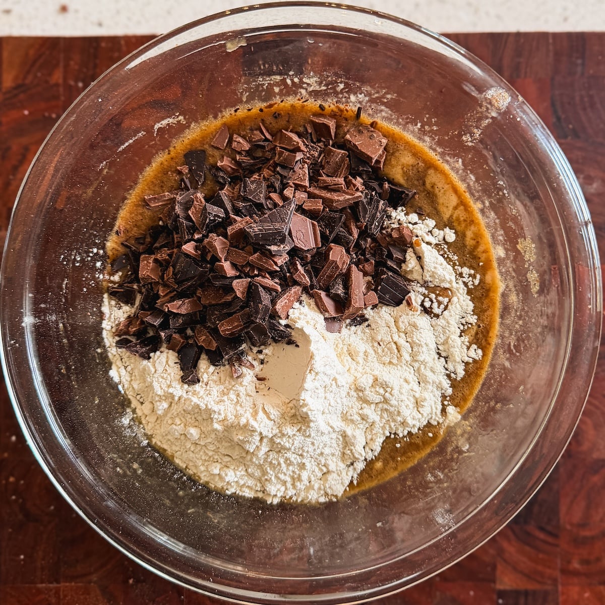 A glass bowl on a wooden surface contains flour, chopped chocolate, and brown batter, ready to be mixed for baking.