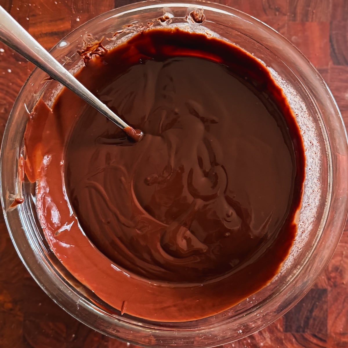 A glass bowl filled with smooth chocolate ganache with a metal spoon resting inside. The bowl is placed on a wooden surface.