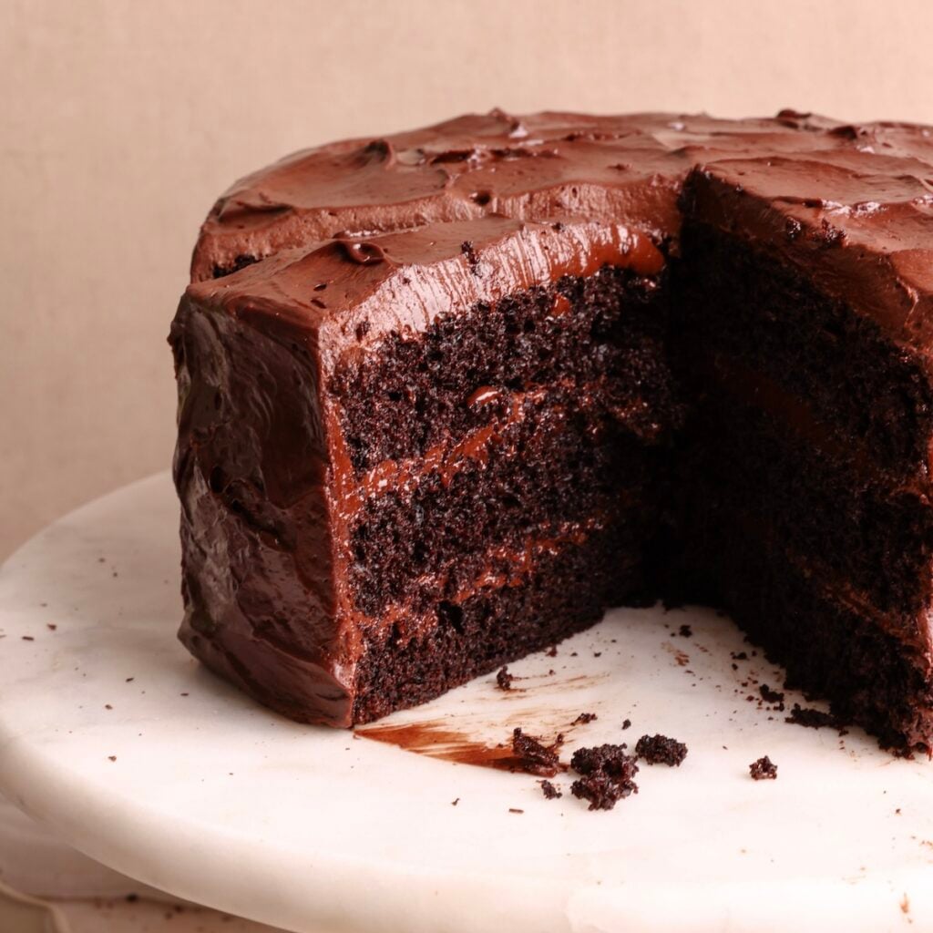 A rich Matilda style chocolate cake with chocolate frosting sits on a marble surface. A large slice has been cut out, revealing moist, dark cake layers and thick frosting inside. Crumbs are scattered near the cut section.