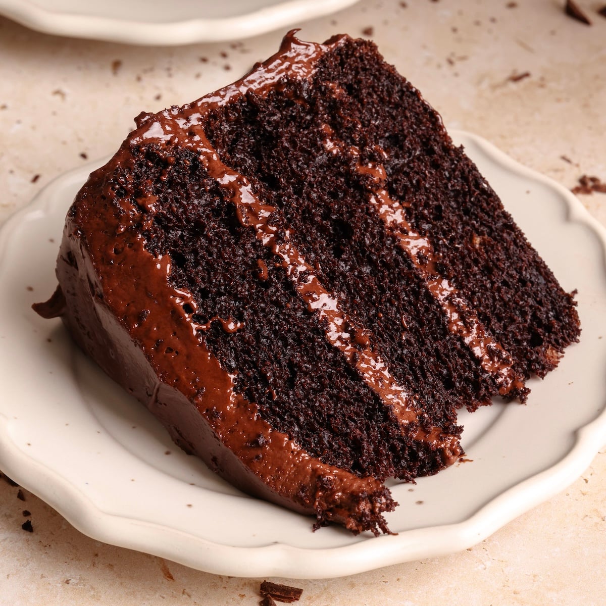 A slice of rich, moist chocolate cake with chocolate frosting between three layers, served on a white scalloped plate.