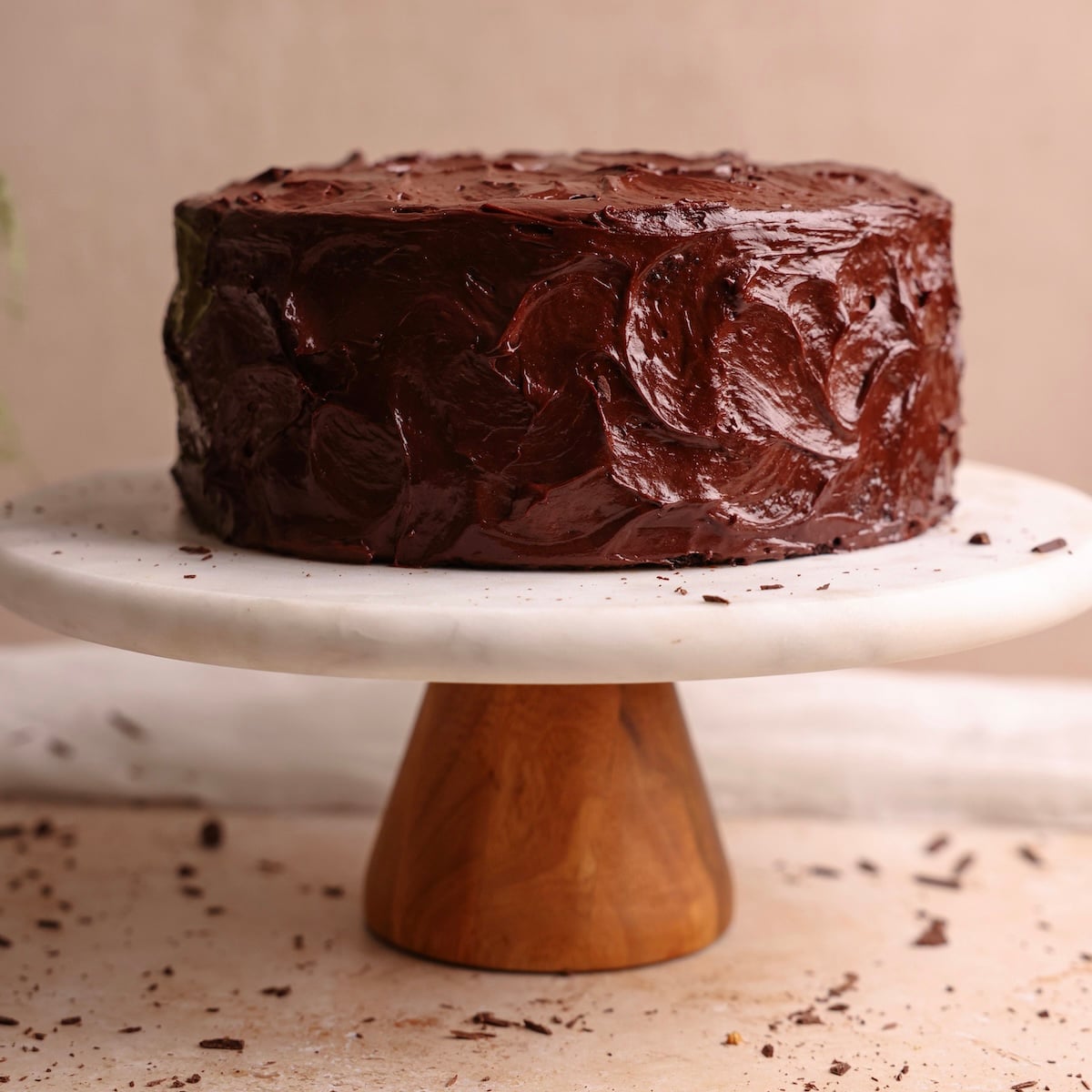 A round chocolate cake with glossy chocolate fudge frosting sits on a marble and wood cake stand, with chocolate crumbs scattered around.