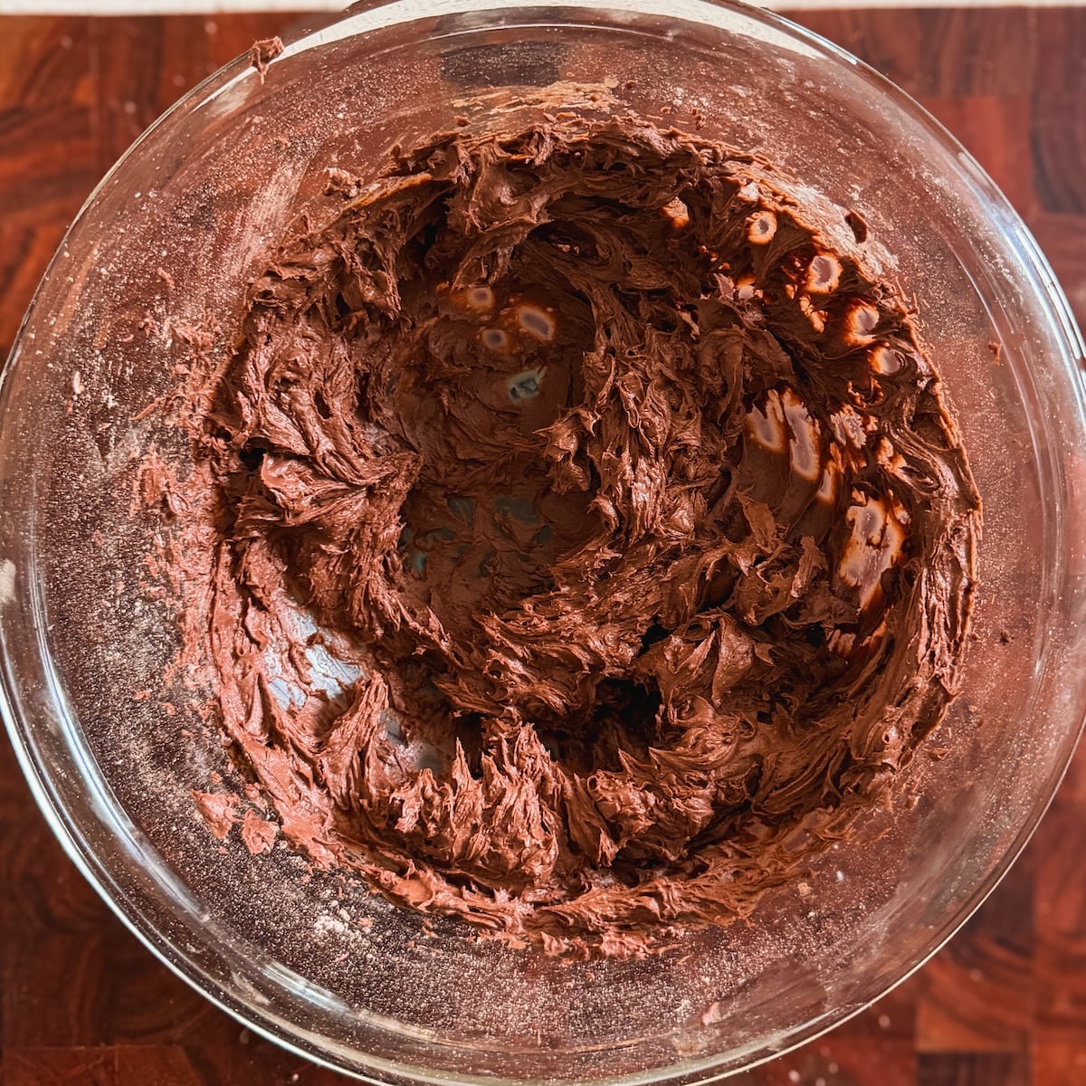 A glass bowl filled with thick, creamy chocolate frosting sits on a wooden surface