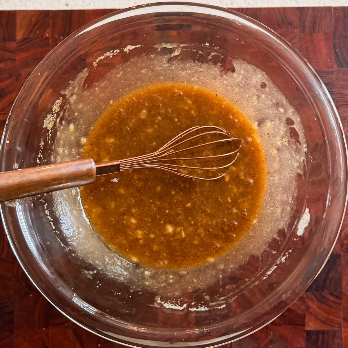 A metal whisk sits in a glass bowl containing a mixture of brown butter, brown sugar and banana, on a wooden countertop.