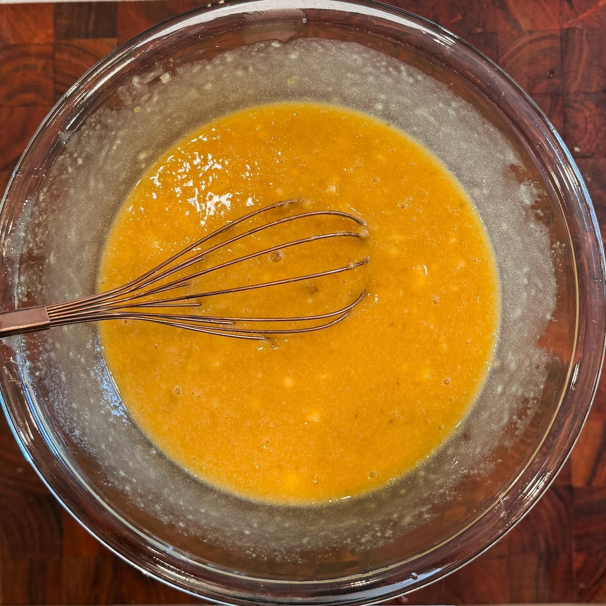 A metal whisk rests in a glass bowl filled with a yellow-orange batter, set on a dark wooden surface.