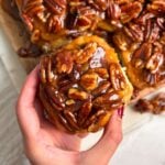A hand holds a sticky bun topped with glossy caramelized pecans, with more pecan-topped buns visible in the background.