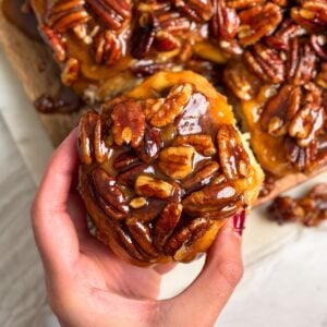 A hand holds a sticky bun topped with glossy caramelized pecans, with more pecan-topped buns visible in the background.