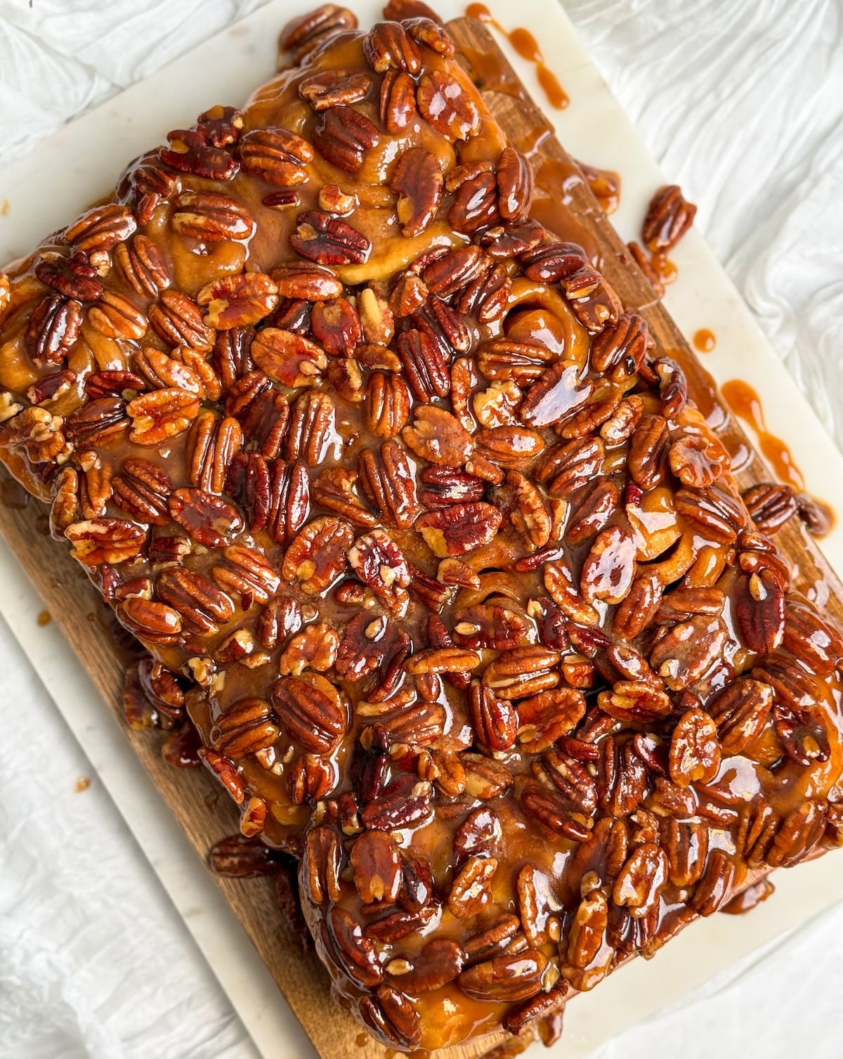Overhead view of a sticky rectangular pecan cinnamon rolls on a wooden board with parchment paper, set on a textured white surface.
