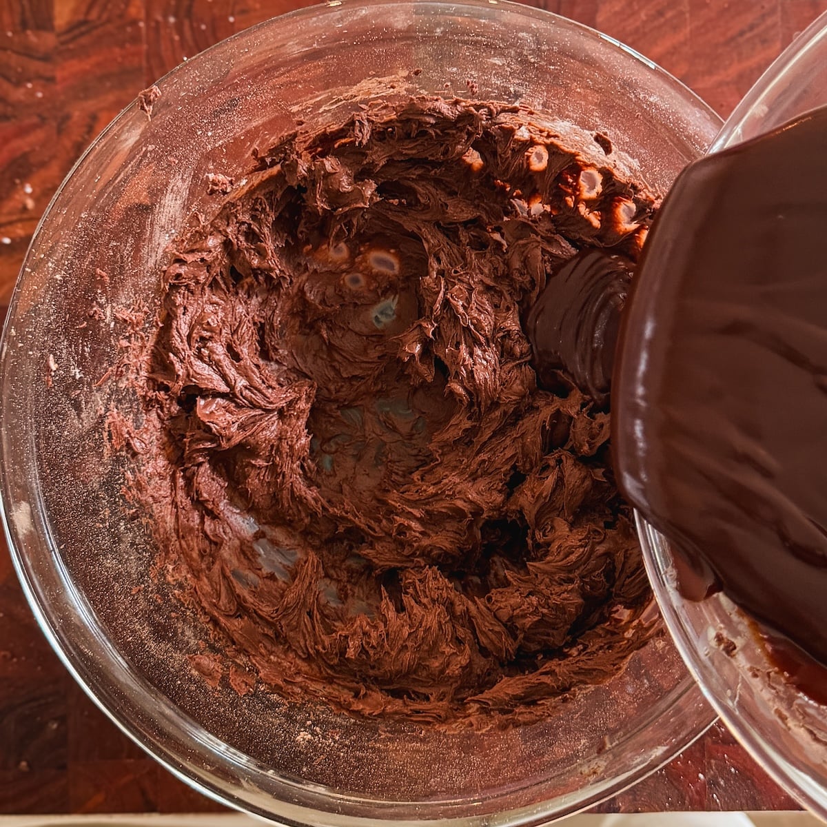 A glass bowl with chocolate frosting being mixed, while more chocolate ganache is being poured in from another container on a wooden surface.