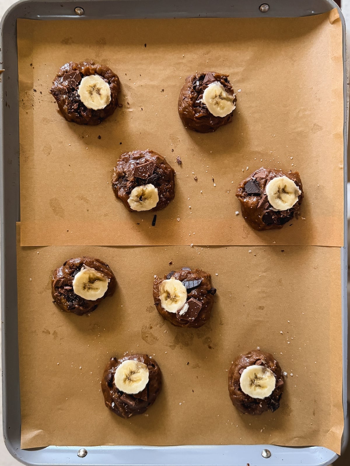 A baking tray with eight banana chocolate cookie dough balls topped with banana slices, placed on brown parchment paper, ready to be baked.
