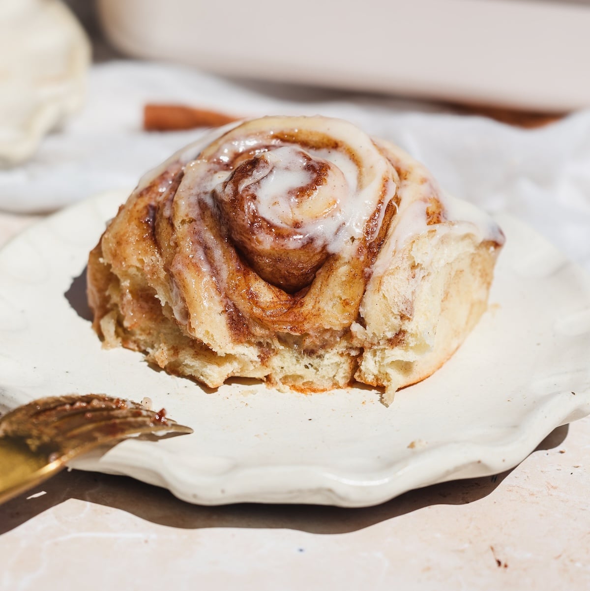 A cinnamon roll topped with icing sits on a white plate with a bite taken out. It looks soft and gooey with a cinnamon filling. A gold fork rests nearby, and the scene is bright and inviting.