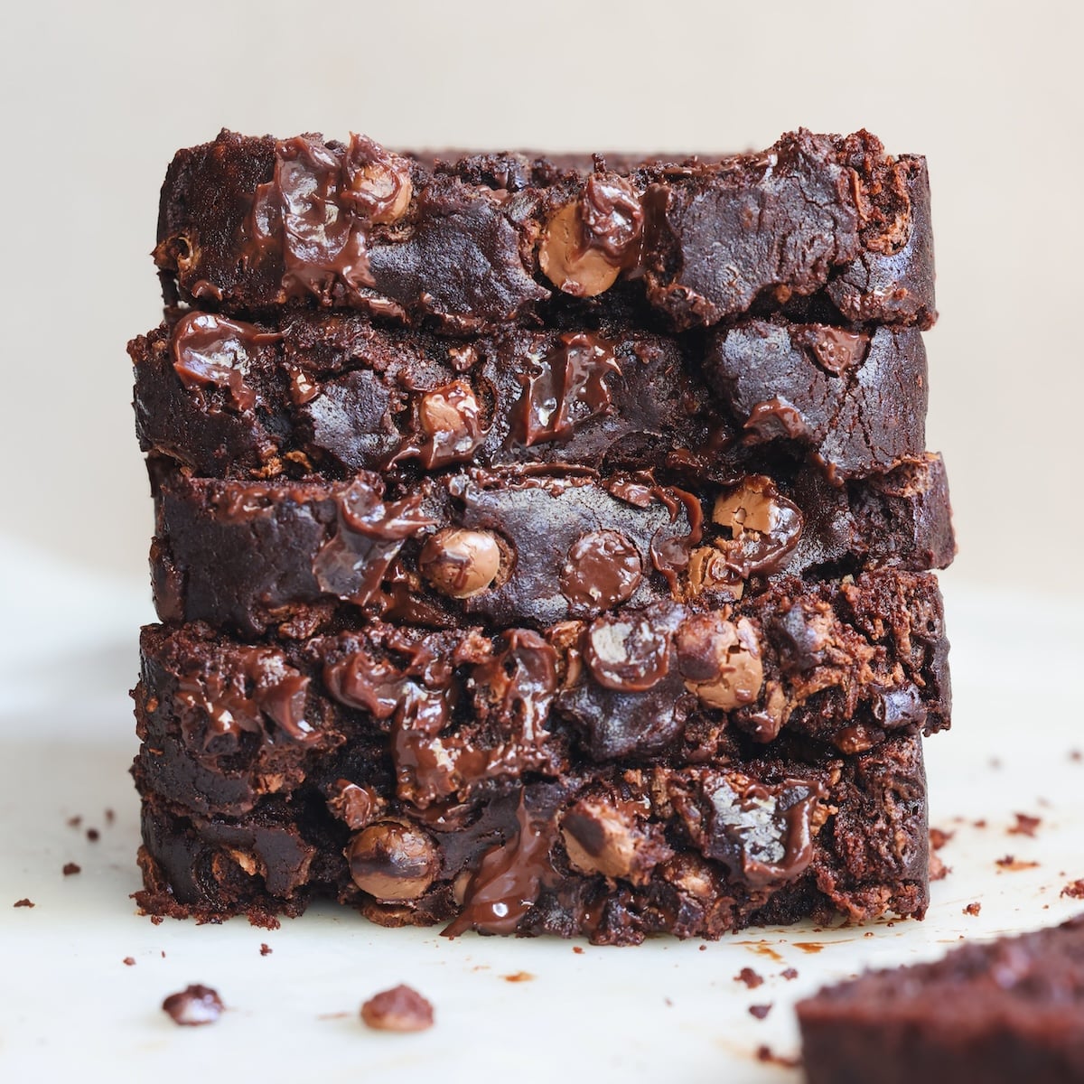 A close-up of four thick, stacked slices of rich, fudgy chocolate banana bread with melted chocolate chips, resting on a light surface with chocolate crumbs scattered around.