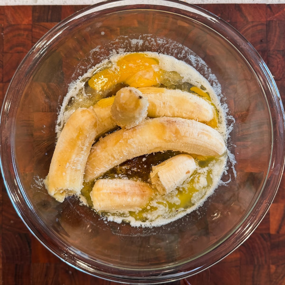 A glass bowl on a wooden surface contains peeled bananas, melted butter, and cracked eggs, ready to be mixed