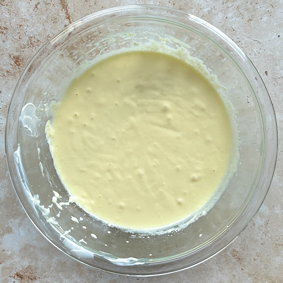 A glass bowl filled with pale yellow mixture of eggs and wet ingredients sits on a light brown countertop. The batter is smooth with a few small bubbles on the surface.