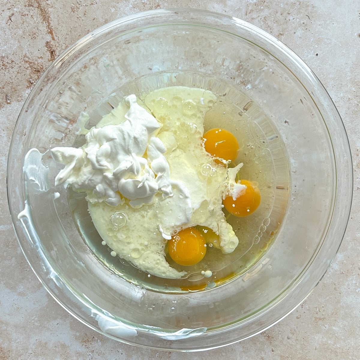 A glass bowl with three raw eggs, a dollop of sour cream, and other creamy ingredients on a beige countertop, ready for mixing.
