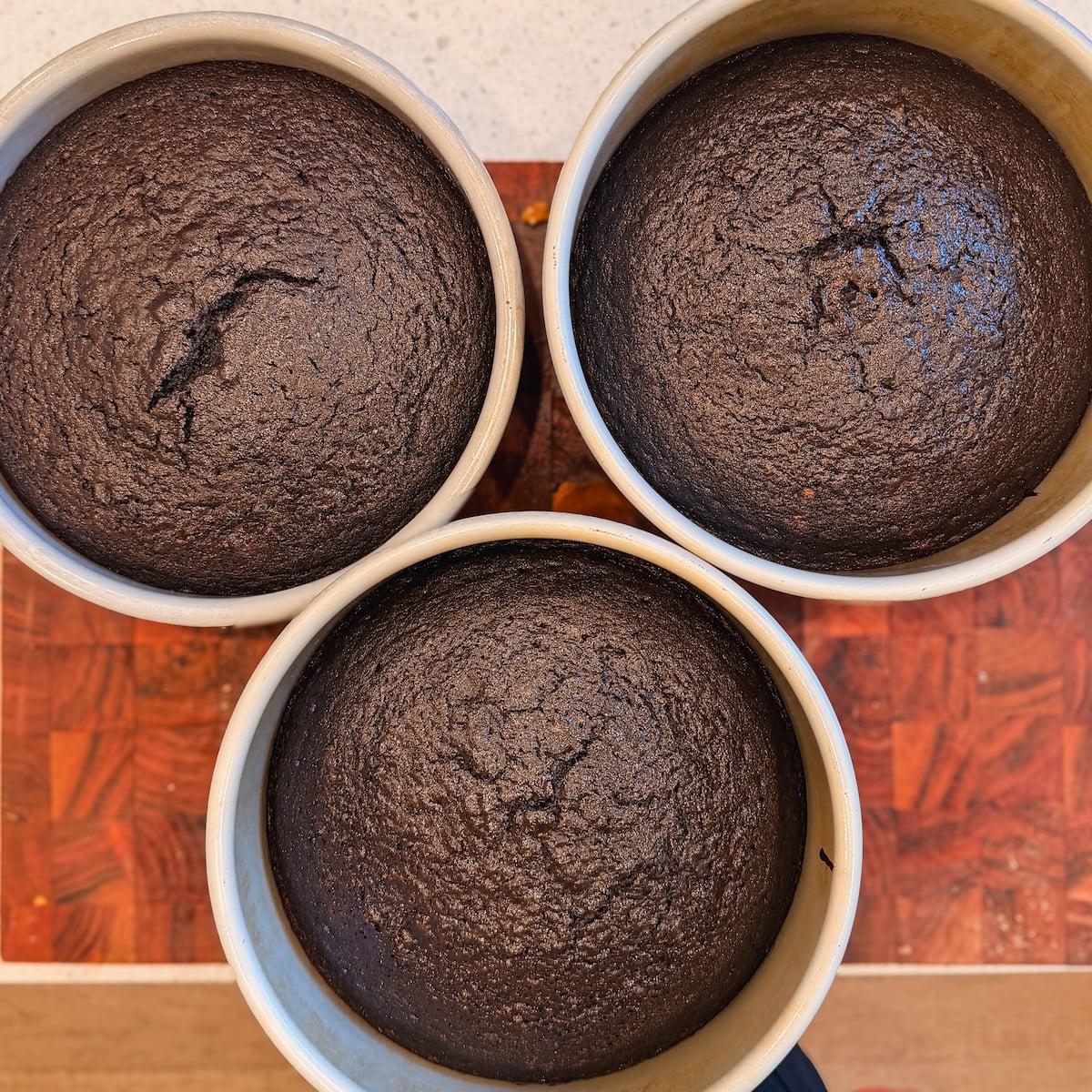 Three round chocolate cakes in baking pans, freshly baked, with cracked tops. The cakes are arranged in a triangular pattern on a wooden surface.