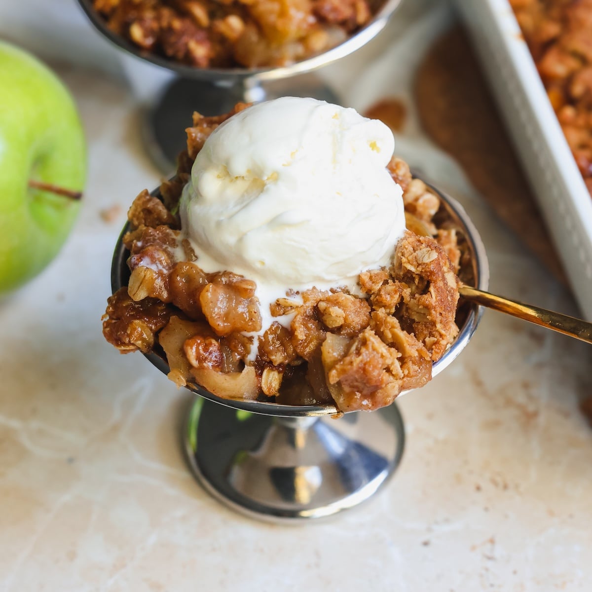 A metal dish filled with apple crisp topped with a scoop of vanilla ice cream, placed on a light-colored surface with a green apple and a baking tray in the background.