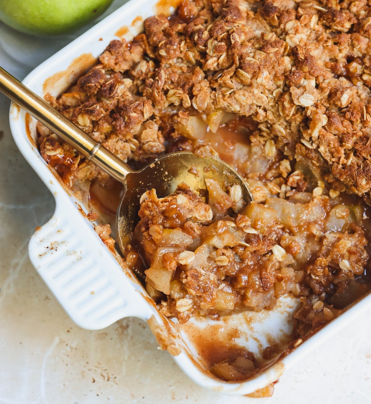 A close-up of a baked apple crisp in a white dish, topped with a golden oat peanut butter crumble. A gold serving spoon rests in the dessert, revealing warm spiced apples inside.
