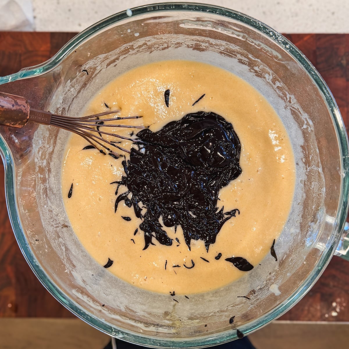 A glass mixing bowl with tan batter and a pool of melted chocolate batter being whisked together on a wooden surface.
