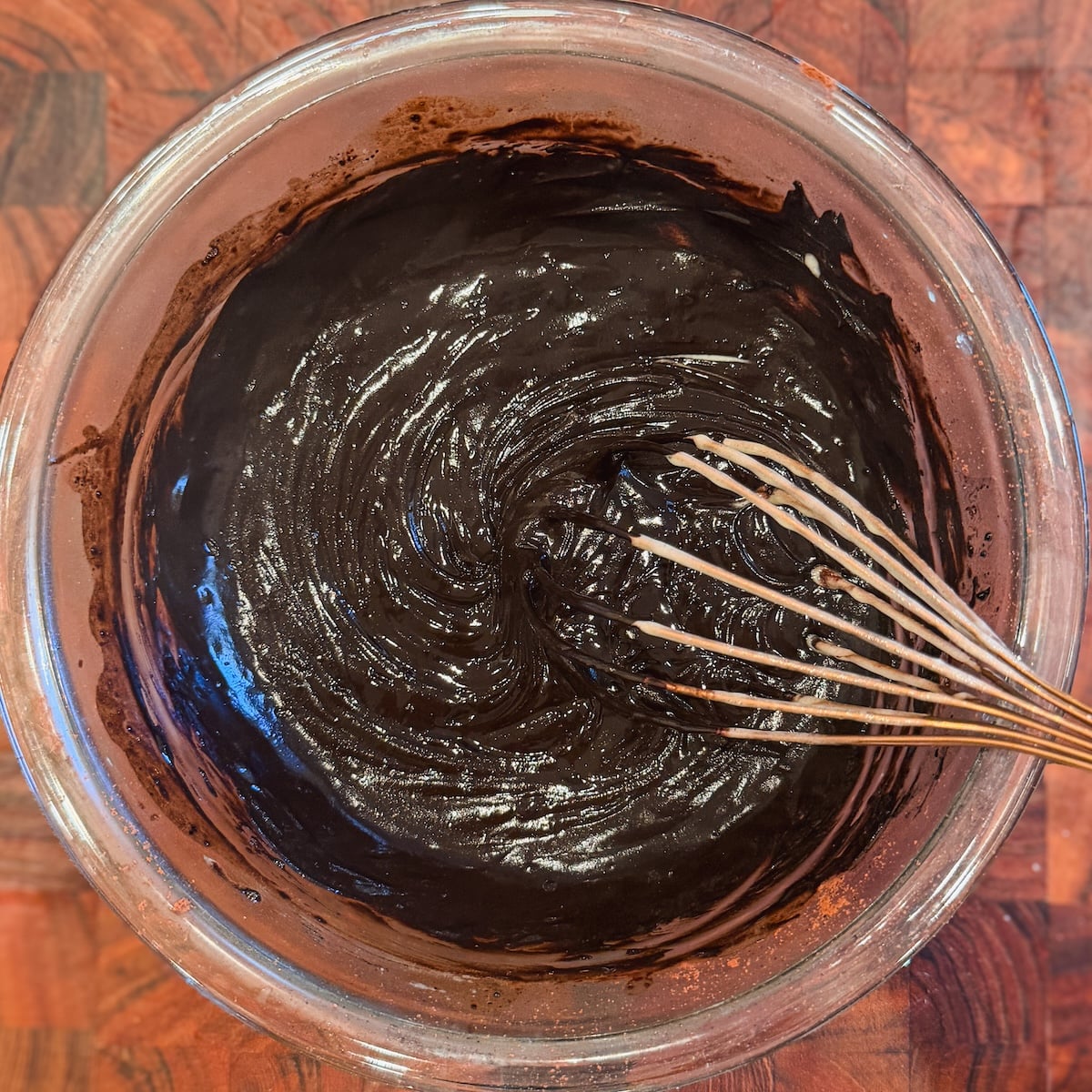 A glass bowl filled with thick, dark chocolate batter being whisked, sitting on a brown wooden surface.