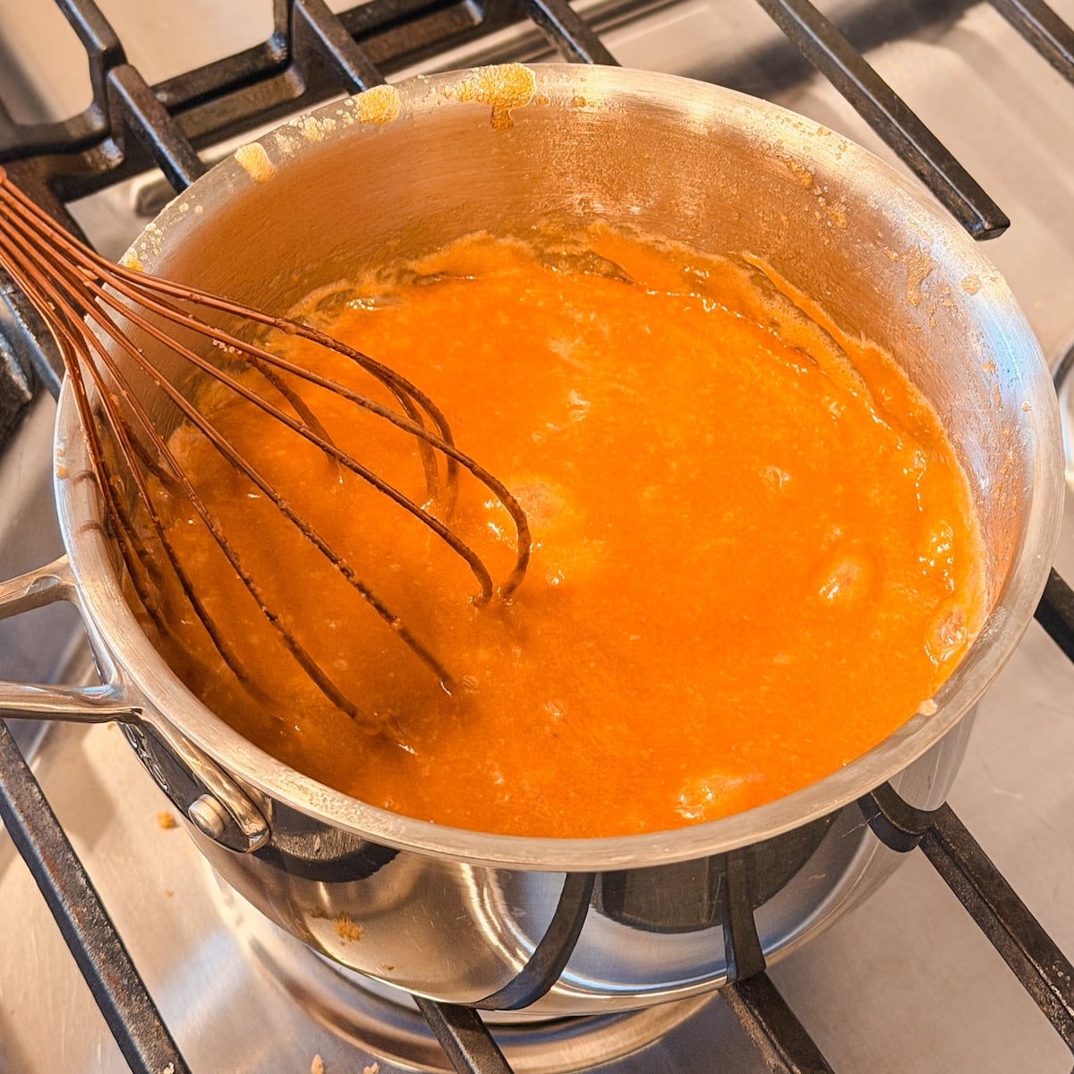 A whisk stirring a pot of bubbly toffee sauceon a stovetop burner.
