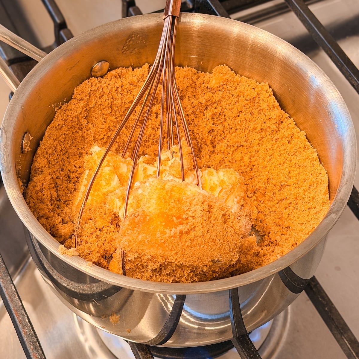 A whisk mixing butter and brown sugar in a stainless steel saucepan on a stovetop, with the mixture beginning to combine and melt.