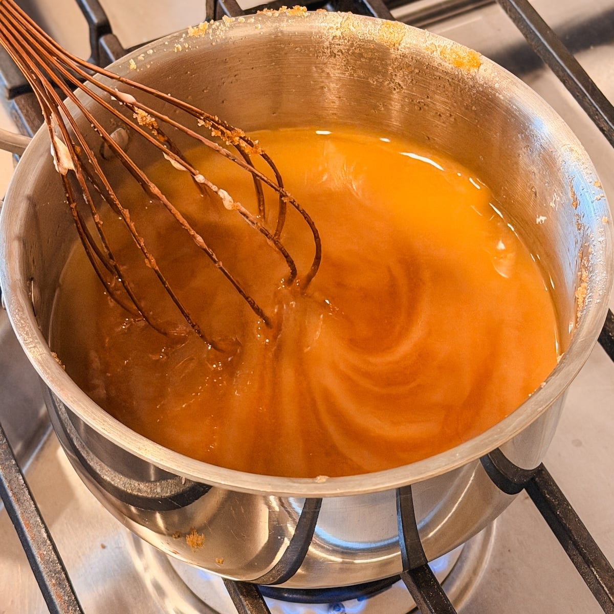 A metal whisk stirs a golden-brown liquid in a stainless steel pot on a gas stove. The mixture appears smooth, and some butter is floating on top